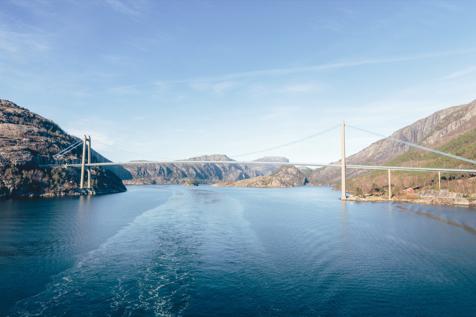 A suspension bridge spanning a fjord between rocky hillsides, seen from the wake of a boat