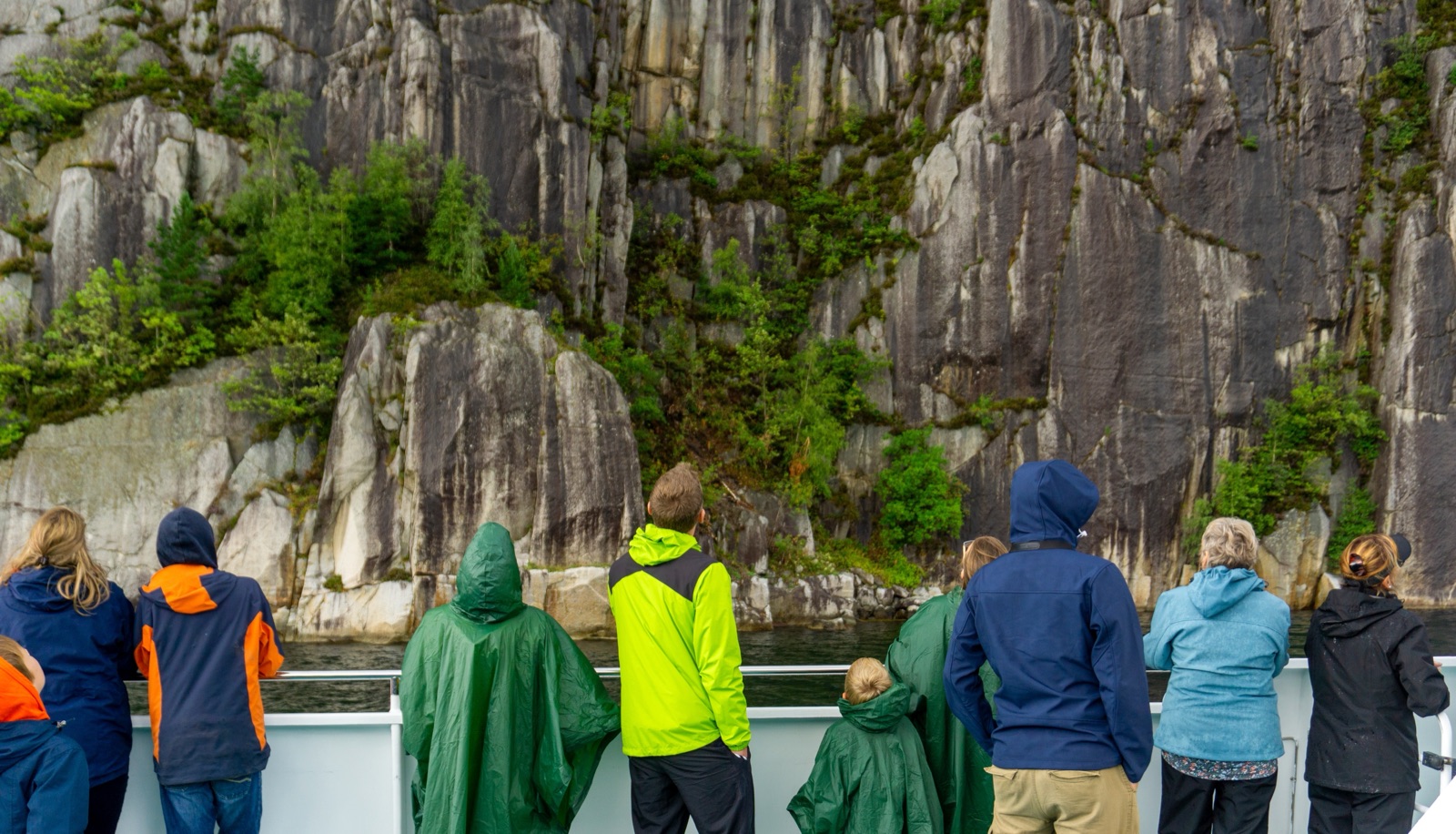 Passengers in rain jackets at the rail of a boat, looking up at a sheer rock face