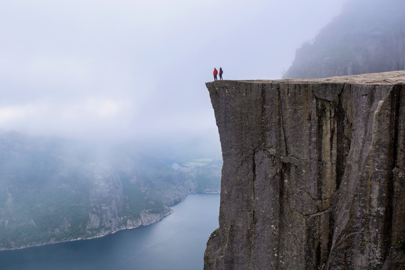 Two hikers at the edge of Preikestolen's flat-topped cliff, with mist over the fjord below